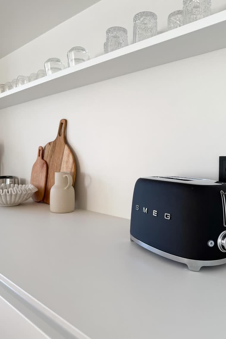 White kitchen counter with a black retro toaster, wood cutting boards and glassware on a floating shelf