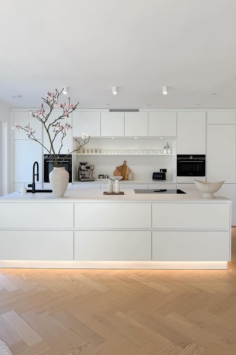 All-white minimalist kitchen with a cherry blossom branch on the island and a herringbone wood floor